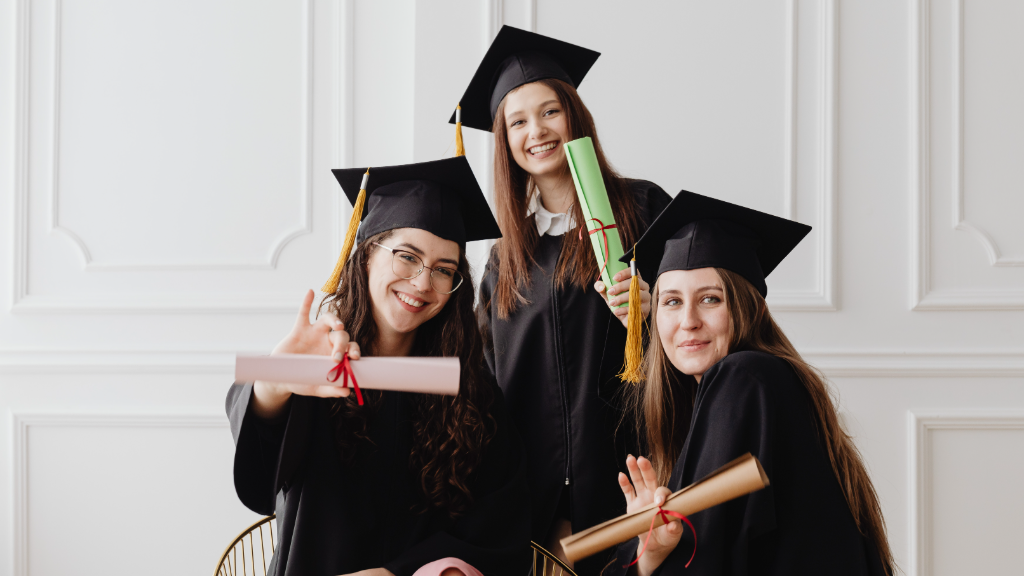 Four graduates in maroon gowns and mortarboards smile together against a white wall